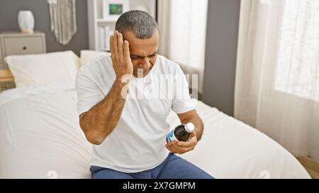 Reifer hispanischer Mann, der Kopfschmerzen hat, während er Medizin in einem Schlafzimmer hält. Stockfoto