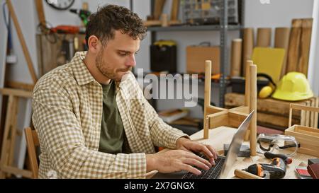 Ein junger Mann mit Bart arbeitet an einem Laptop in einer Tischlerei, umgeben von Werkzeugen und Holzstücken. Stockfoto