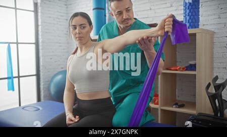 Eine Frau unterzieht sich einer Schulterrehabilitation mit einem männlichen Physiotherapeuten mit einem violetten Widerstandsband in einer hell beleuchteten Physiotherapieklinik. Stockfoto