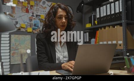 Eine konzentrierte Detektivin untersucht einen Fall in ihrem Büro, umgeben von Beweisen, mit einem Laptop. Stockfoto