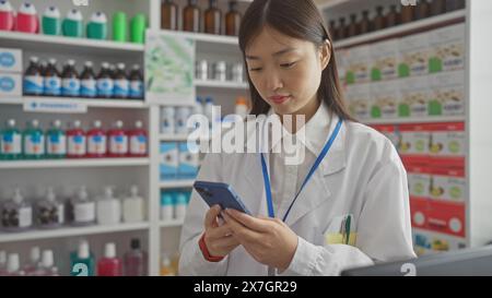 Eine junge asiatische Apothekerin benutzt ein Smartphone in einer modernen Apotheke, die mit medizinischen Vorräten gefüllt ist. Stockfoto
