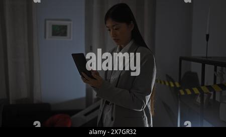 Eine asiatische Frau analysiert einen Tatort in einem schwach beleuchteten Wohnzimmer und nimmt ihre Beobachtungen mit einem Tablet auf. Stockfoto