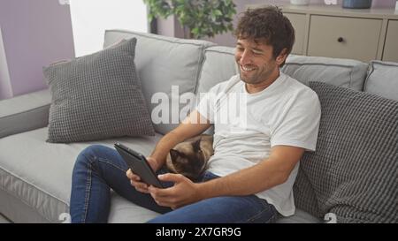 Junger Mann lächelt, während er Tablette benutzt und mit der Katze drinnen auf der Couch kuschelt Stockfoto