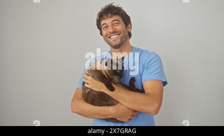 Ein lächelnder junger Mann in einem blauen Hemd hält liebevoll eine siamesische Katze vor weißem Hintergrund. Stockfoto