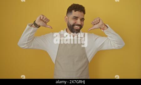 Ein fröhlicher hispanischer Mann in einer Schürze auf gelbem Hintergrund, der stolz auf sich selbst zeigt. Stockfoto