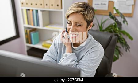 Lächelnde hispanische Frau mit kurzen blonden Haaren, die am Telefon in einer modernen Büroumgebung spricht. Stockfoto