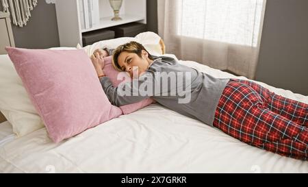 Hispanische Frau mit kurzen blonden Haaren, die im Schlafanzug auf dem Bett liegt und entspannt in einer gemütlichen Schlafzimmerumgebung aussieht. Stockfoto