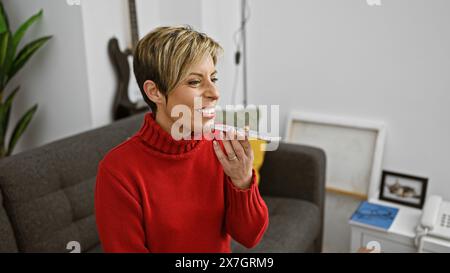 Lächelnde hispanische Frau mit kurzen blonden Haaren, die zu Hause eine Sprachnachricht sendet. Stockfoto