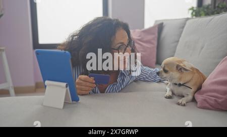 Eine Frau mittleren Alters interagiert mit einem chihuahua auf einem Sofa, während sie eine Kreditkarte in der Hand hat und schlägt einen Online-Kauf zu Hause vor. Stockfoto