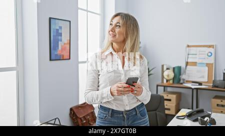 Eine professionelle kaukasierin in einem modernen Büro hält ein Smartphone und blickt nachdenklich weg. Stockfoto