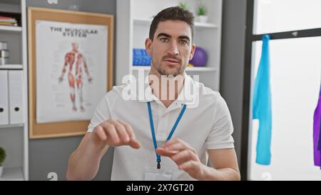 Gutaussehender hispanischer Therapeut in einer Rehabilitationsklinik, trägt ein Schlüsselband und posiert drinnen mit einem anatomischen Poster im Hintergrund. Stockfoto