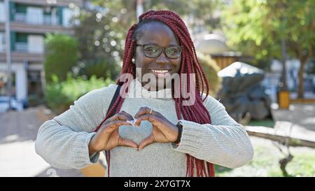 Eine fröhliche afrikanerin mit Zöpfen, die draußen in der Stadt ein Herzzeichen bilden. Stockfoto