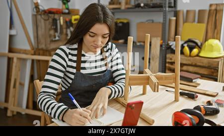 Die junge hispanische Frau macht Notizen in einer Tischlerei mit Werkzeugen Stockfoto