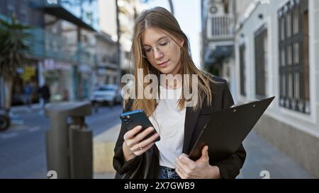 Eine junge blonde Frau in Geschäftskleidung überprüft ihr Handy, während sie ein Klemmbrett in einer belebten Straße hält. Stockfoto