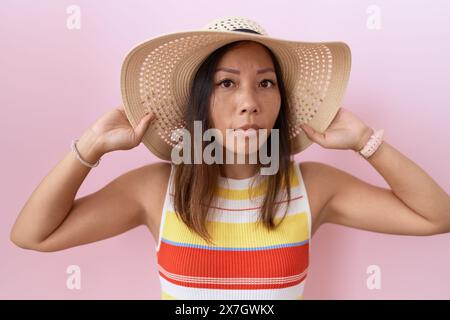 chinesin mittleren Alters mit Sommerhut über rosa Hintergrund, ahnungsloser und verwirrter Ausdruck. Zweifel. Stockfoto