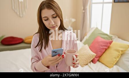 Eine junge Frau im pinkfarbenen Pyjama hält einen Becher und ein Smartphone in gemütlicher Schlafzimmerumgebung. Stockfoto