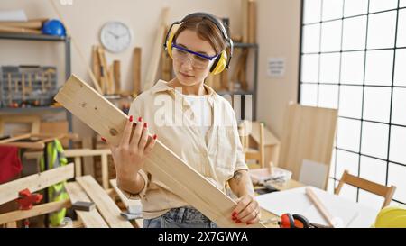 Eine fokussierte Frau mit Schutzkleidung untersucht eine Holzdiele in einer gut ausgestatteten Tischlerei. Stockfoto