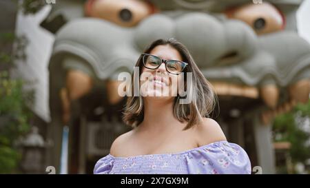 Fröhlich schöne hispanische Frau mit Brille posiert mit selbstbewusstem, freudigem Lächeln bei namba yasaka jinja in osaka, stehend neben dem beeindruckenden Riesen dra Stockfoto