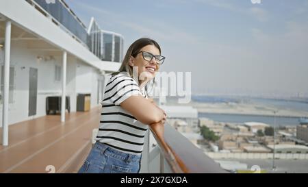 Lächelnde junge Frau, die sich auf ein Kreuzfahrtschiff lehnt, das den Ozean überblickt, mit gestreiftem Hemd und Brille. Stockfoto