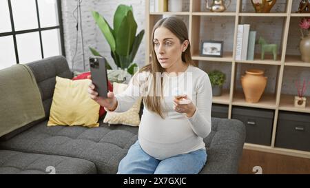 Eine junge schwangere hispanische Frau zu Hause, die eine Videoberatung mit Smartphone und Medikamentenflasche im Wohnzimmer hat. Stockfoto