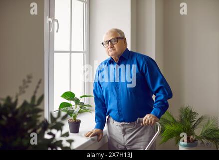Senior Mann, der zu Hause in der Rehabilitation am Fenster stand, mit seiner Krücken und in die Kamera schaute. Stockfoto