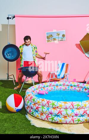 Überfröhlicher Mann im farbenfrohen, tropischen Hemd-Kochen-Grill, Würstchen im Studio mit rosa Hintergrund und Kinderpool. Stockfoto
