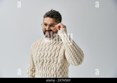 Ein bärtiger Mann verströmt Charme in einem weißen Pullover und einer Brille vor einem grauen Studio-Hintergrund. Stockfoto