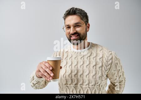 Bärtiger Mann in weißem Pullover hält eine dampfende Kaffeetasse in einem gemütlichen Studio-Setting auf grauem Hintergrund. Stockfoto