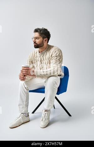 Ein bärtiger Mann in einem weißen Pullover sitzt auf einem blauen Stuhl und kreuzt seine Arme in besinnlicher Pose vor grauem Hintergrund. Stockfoto