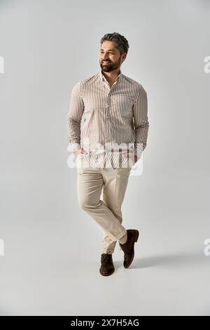 Ein stilvoller Mann mit Bart posiert in eleganter Kleidung und steht mit Händen in Taschen auf einem grauen Studiohintergrund. Stockfoto