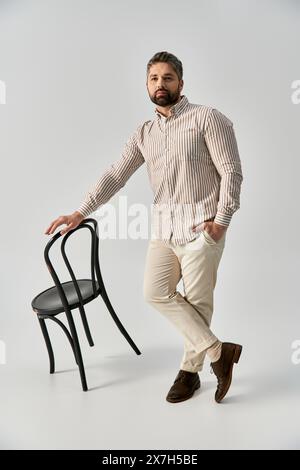 Ein bärtiger Mann in eleganter Kleidung, der neben einem eleganten schwarzen Stuhl vor einem grauen Studiohintergrund posiert. Stockfoto