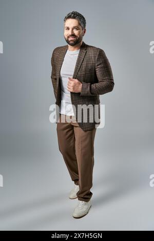 Ein fesselnder Mann mit Bart posiert in einem eleganten braunen Anzug und weißem Hemd vor grauem Studio-Hintergrund. Stockfoto