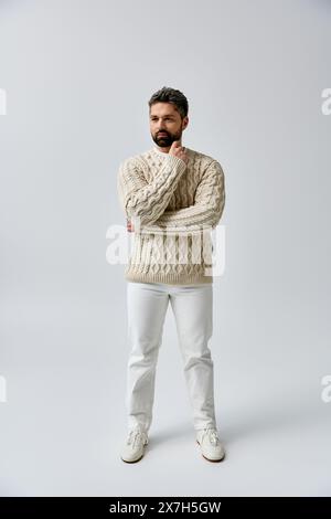 Ein bärtiger Mann strahlt Eleganz in einem weißen Pullover und einer Hose vor einem grauen Studio-Hintergrund aus. Stockfoto