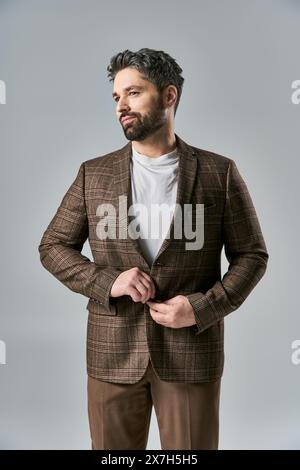 Ein charismatischer Mann mit Bart in brauner Jacke und Hose posiert elegant vor grauem Studio-Hintergrund. Stockfoto