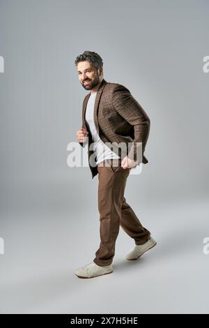 Ein bärtiger Mann posiert in einer eleganten braunen Jacke und einem knusprigen weißen Hemd vor grauem Hintergrund in einem Studio-Setting. Stockfoto