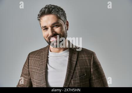 Ein bärtiger Mann strahlt Charme und Eleganz in seiner modischen Jacke vor grauem Hintergrund aus. Stockfoto