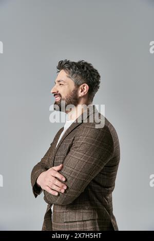 Ein raffinierter Mann mit Bart, in eleganter Kleidung gekleidet, steht mit überkreuzten Armen vor grauem Hintergrund. Stockfoto