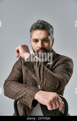 Ein bärtiger Mann in einer braunen Jacke strahlt Stil aus, während er eine schwarze Tasche vor grauem Hintergrund hält. Stockfoto