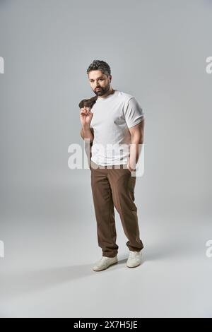 Ein stilvoller Mann mit Bart steht selbstbewusst mit der Hand in der Tasche, gekleidet in eleganter Kleidung, vor grauem Hintergrund in einem Studio. Stockfoto