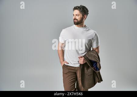 Ein stilvoller Mann mit Bart posiert selbstbewusst in elegantem weißem T-Shirt und brauner Hose vor grauem Studiohintergrund. Stockfoto