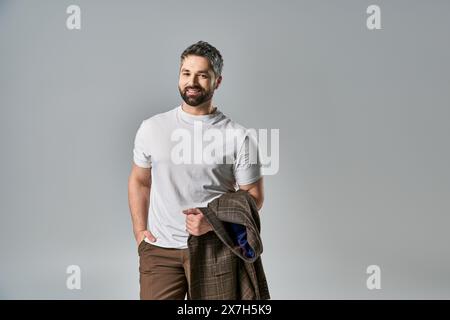 Ein charismatischer Mann mit Bart posiert in eleganter Kleidung vor grauem Atelierhintergrund. Stockfoto