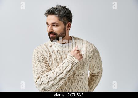 Ein charismatischer Mann mit Bart posiert in einem weißen Pullover vor grauem Studio-Hintergrund. Stockfoto