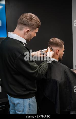 Der Friseur rasiert männliche Schläfen mit Trimmer in der Nähe des Friseurs. Professioneller Friseur schneidet Männerhaare mit einer Maschine im Friseursalon Stockfoto