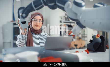 Modernes Büro: Porträt einer muslimischen Geschäftsfrau, die Hijab trägt und an Engineering Project arbeitet, Codierung auf Laptop und veränderte Roboterhandposition. Befähigter Techniker Mit Startup-Projekt. Stockfoto