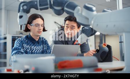 Teamarbeit in multiethnischen Büros: Der Projektmanager unterstützt seinen Kollegen, der an einem Laptop arbeitet. Profis erstellen Programm für Roboterarm. Menschen, Die Automatisierte Robotersysteme Entwickeln. Stockfoto