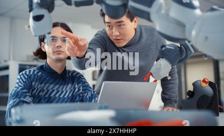 Teamwork in diversen Büros: Smiling Project Manager unterstützt seinen Kollegen, der an einem Laptop arbeitet. Profis erstellen Programm für Roboterarm. Menschen, Die Automatisierte Robotersysteme Entwickeln. Stockfoto