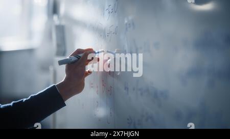 Makroaufnahme eines blauen Markierstifts, der mit einer Hand gehalten wird. Lehrer schreibt Gleichungen auf einem Whiteboard mit mathematischen Formeln. Hochschulbildung an der Technischen Universität Konzept. Stockfoto