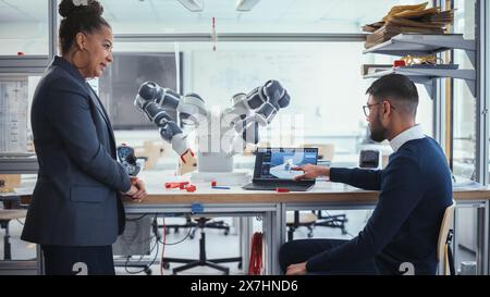 Schwarzer Chefingenieur diskutiert Futuristic Robotic Arm mit einem jungen vielversprechenden männlichen Computerwissenschaftler mit Laptop im Forschungslabor. Innovatives Startup Company-Konzept. Mittlere Aufnahme. Stockfoto