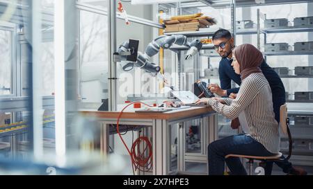 Black Male Engineer hebt Roboterarm mit Controller an, während weibliche Entwicklerin in Hijab Code auf Laptop schreibt. Menschen Entwickeln Innovative Anwendungen. Konzept der High-Tech- und Robotik-Industrie. Stockfoto