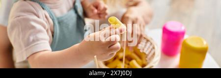 Ein Kleinkind mit lockigen Haaren spielt fröhlich mit trockenen Nudeln und erforscht Texturen und Formen in einem von Montessori inspirierten Lernmoment. Stockfoto